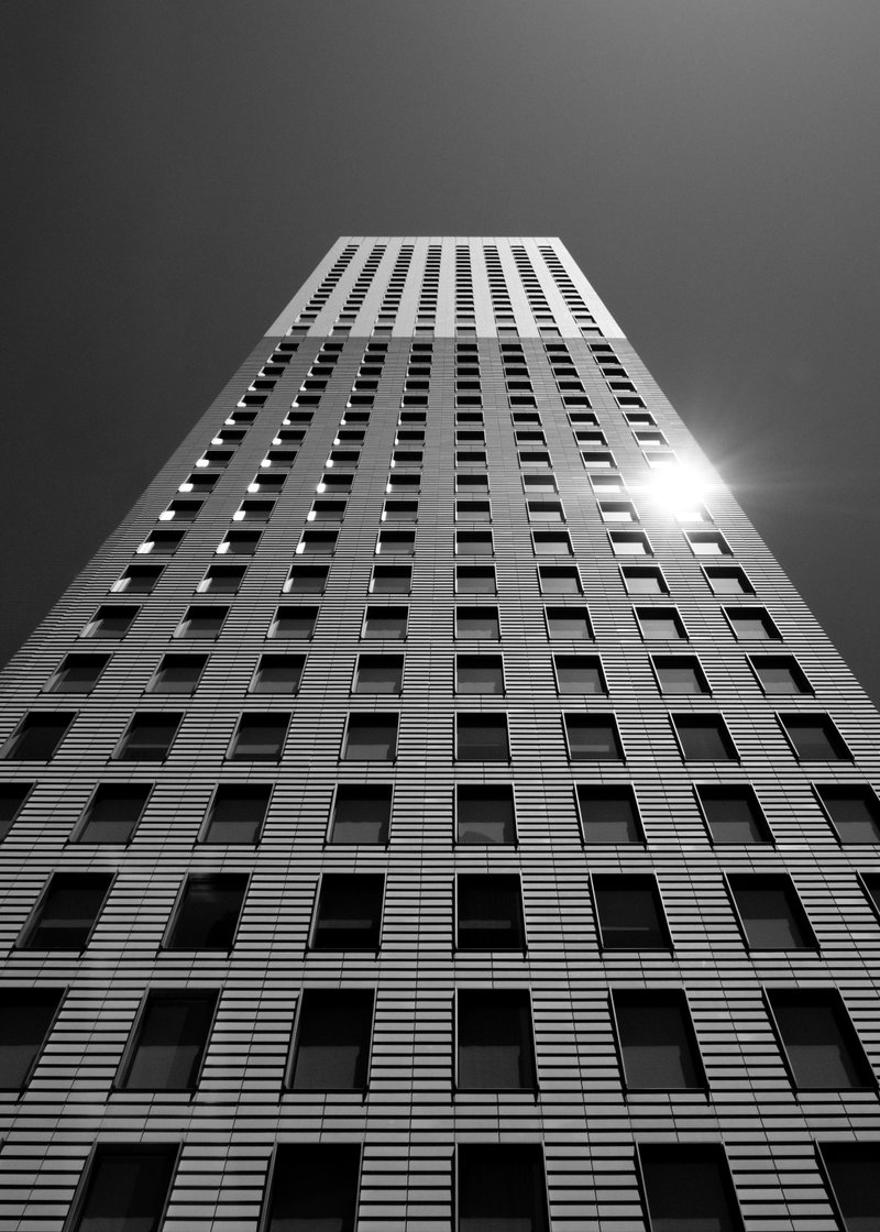 A tall, modern skyscraper with a grid of windows on its facade, shot from a low angle with the sun reflecting off the building's surface, in black and white.