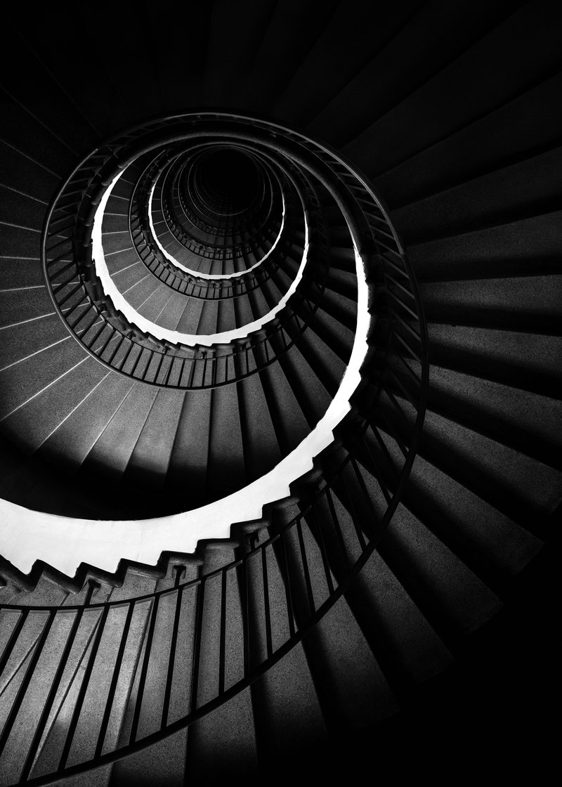 Black and white photo of a spiral staircase looking downward, with railings and steps creating a tunnel effect.