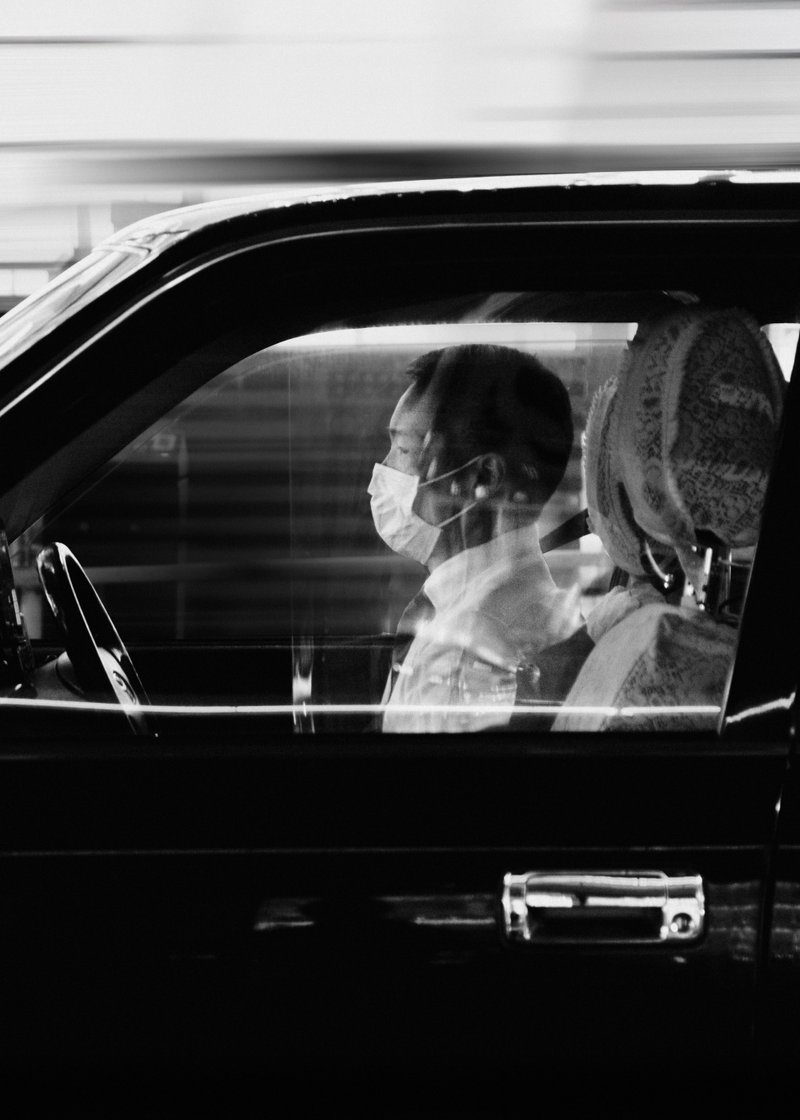 A person wearing a face mask sitting in the driver's seat of a car, seen through the window.