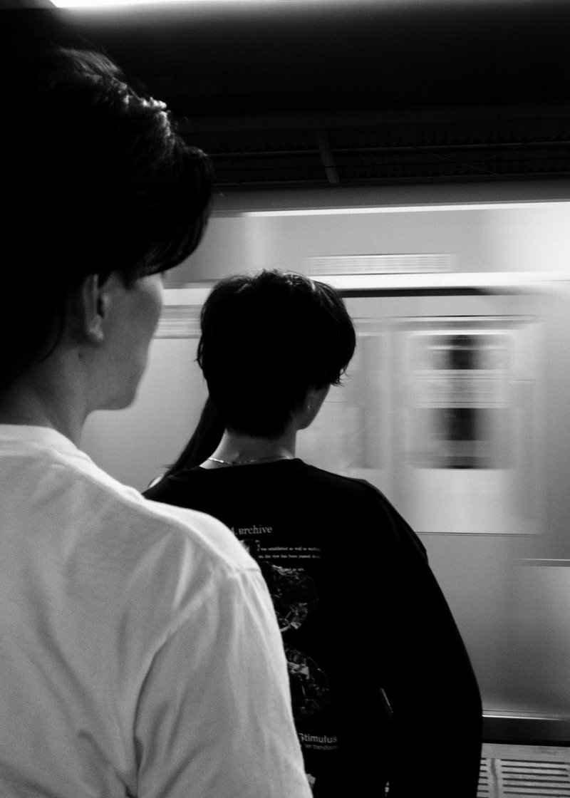 Two people standing with their backs to the camera on a subway platform as a train rushes past in a blur, in black and white.