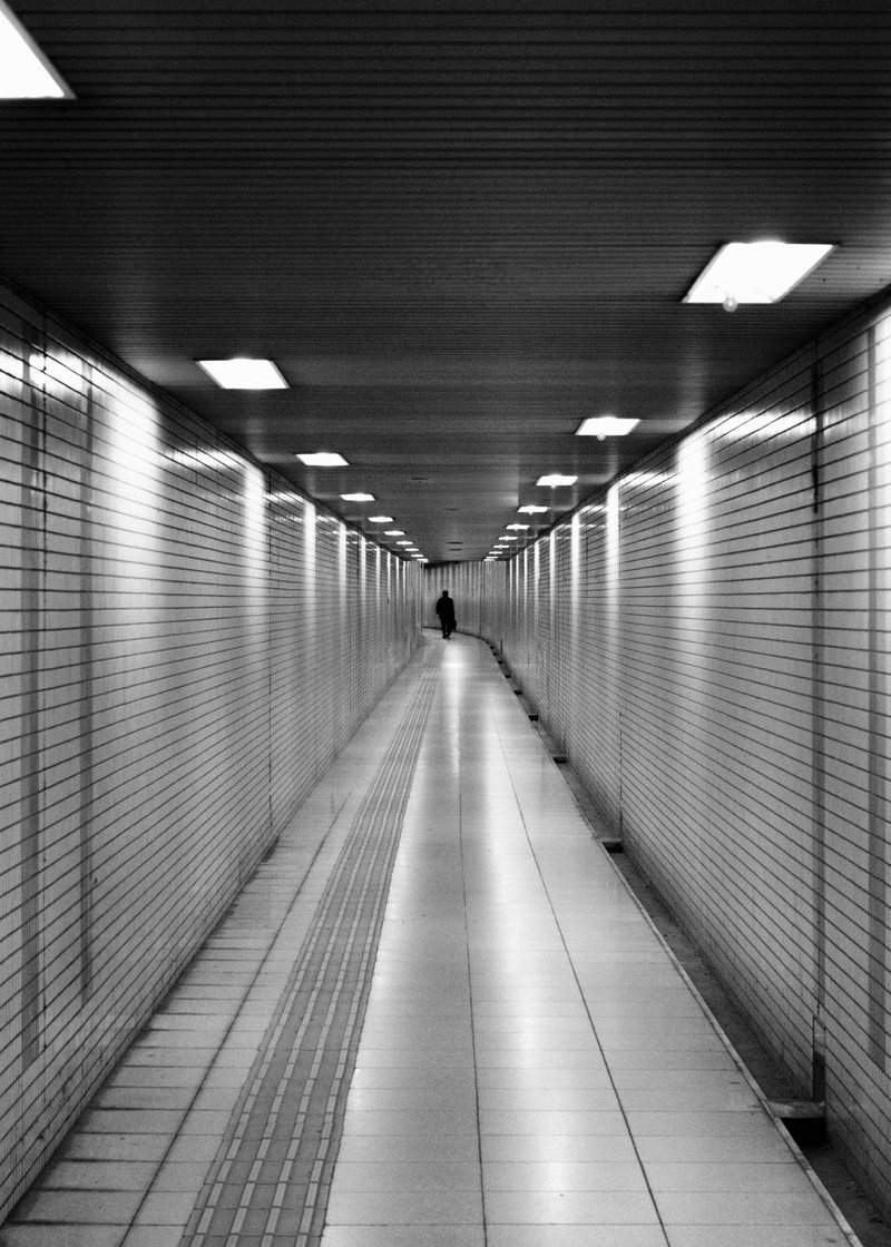 A long, empty underground pedestrian tunnel with tiled walls and ceiling, illuminated by fluorescent lights, with a single person walking away in the distance.