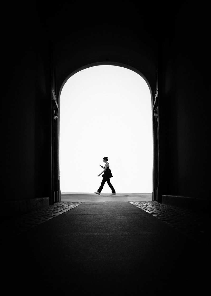 Silhouette of a woman walking while looking at her phone, seen through a dark archway with bright background.