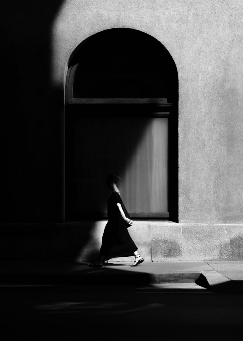 A woman walking past a large arched window on a building, with sharp shadow patterns on the wall, captured in black and white.