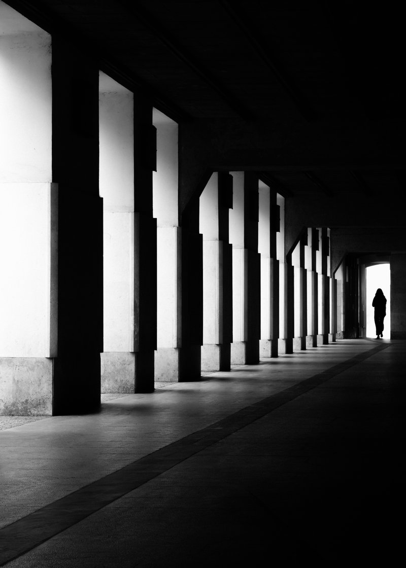 Silhouette of a person walking through an arched corridor with tall columns, in black and white.