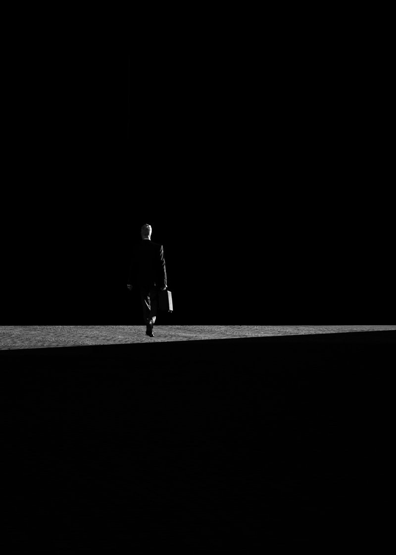 A black-and-white photo of a man in a suit walking away, carrying a briefcase, against a dark background.