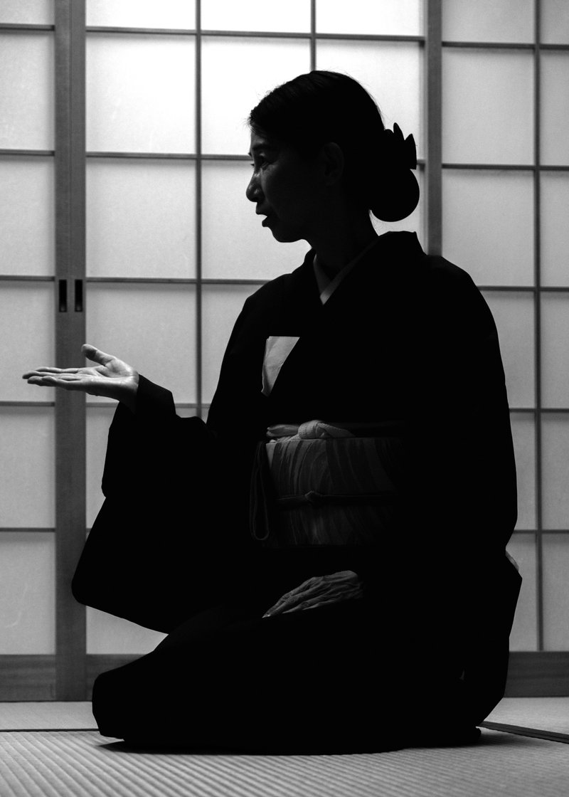 Silhouette of a woman in a kimono seated on a tatami floor in profile, one hand raised, against a backlit shoji screen, in black and white.