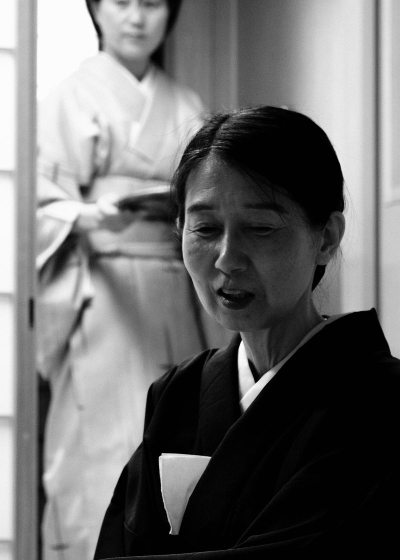 Black and white photo of two women, one in traditional Japanese attire in the foreground with her eyes closed, and another in a nurse outfit standing behind her.