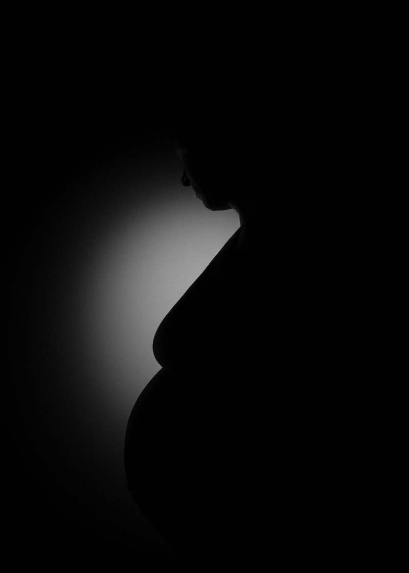 Silhouette of a pregnant woman in profile, with a backlight highlighting her pregnant belly, set against a dark background.