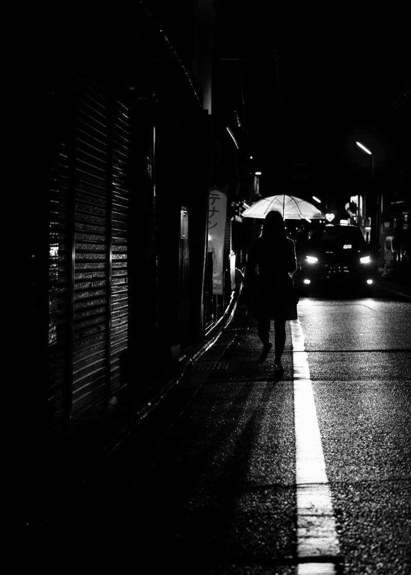 A silhouette of a woman walking on a wet city street at night holding an umbrella, with a car approaching in the background.
