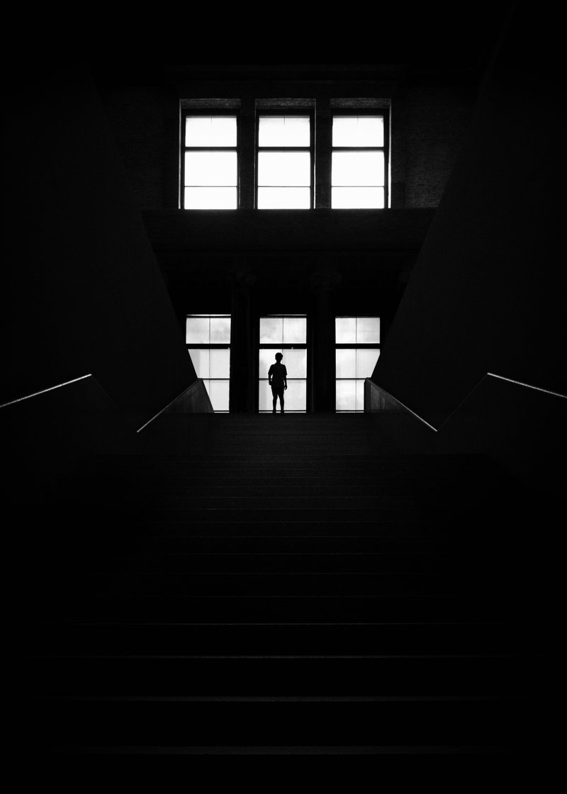 Silhouette of a person standing at the top of a staircase inside a building with large windows, viewed in black and white.