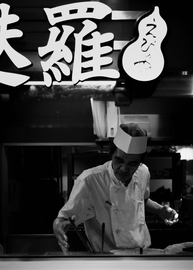 A chef wearing a paper hat and white uniform working in a kitchen, seen through a glass window with Asian characters above.
