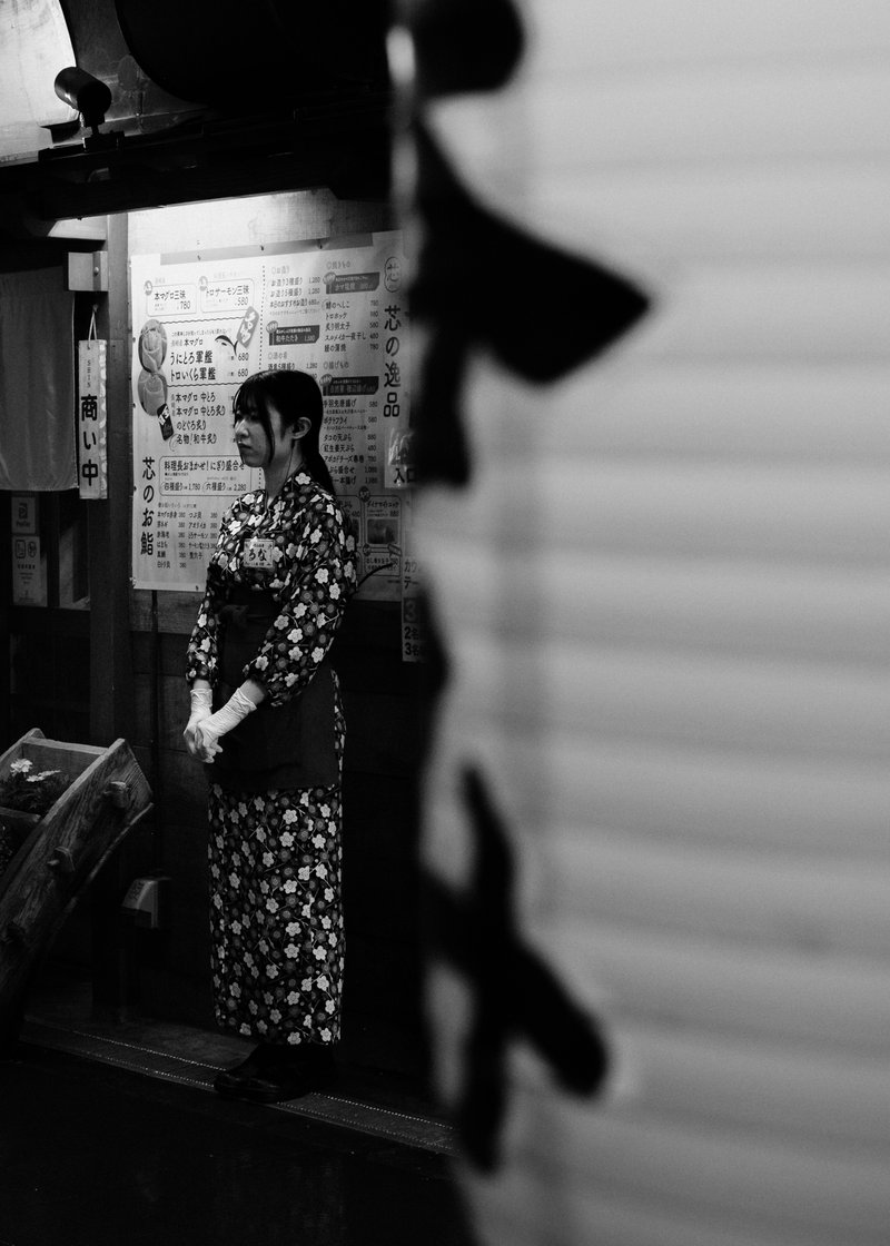 A woman in a floral kimono and apron standing at the entrance of a Japanese restaurant, with a menu board behind her, in black and white.