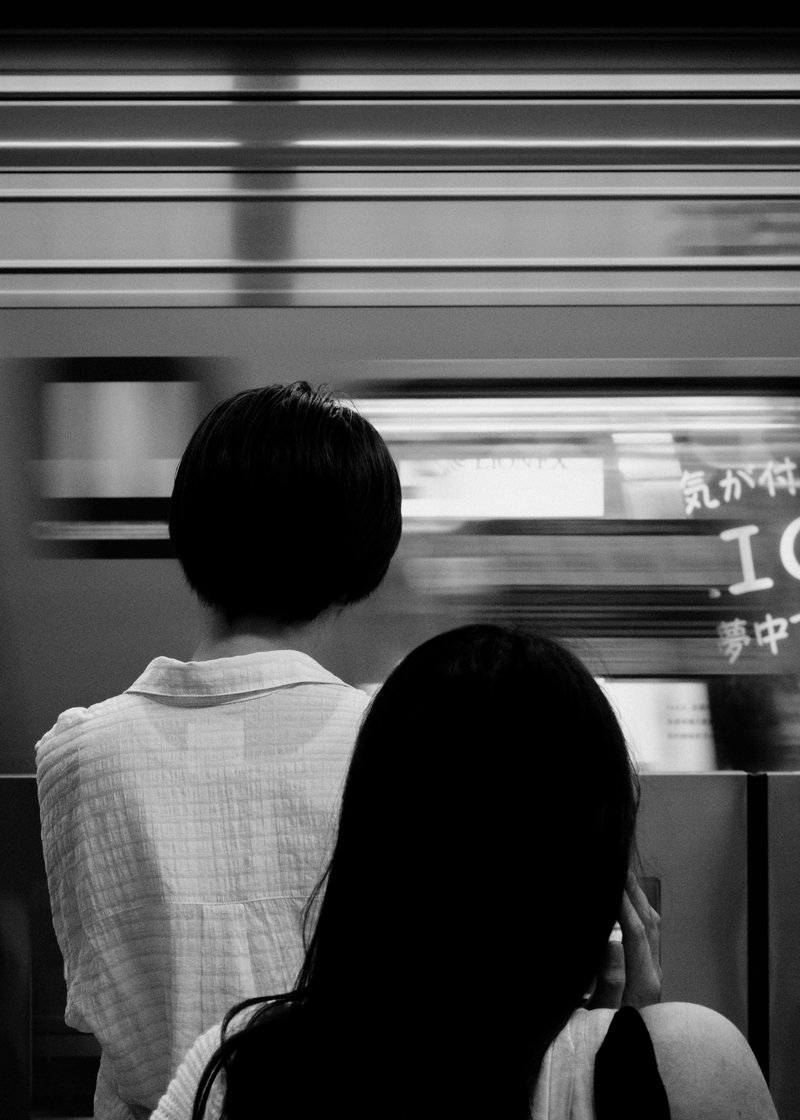 A black and white photo of two people with their backs to the camera, waiting near a moving train.