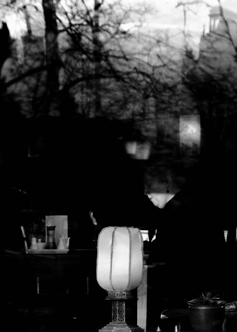 Black and white photo of a woman sitting at a table through a glass window reflecting tree branches and a cloudy sky.