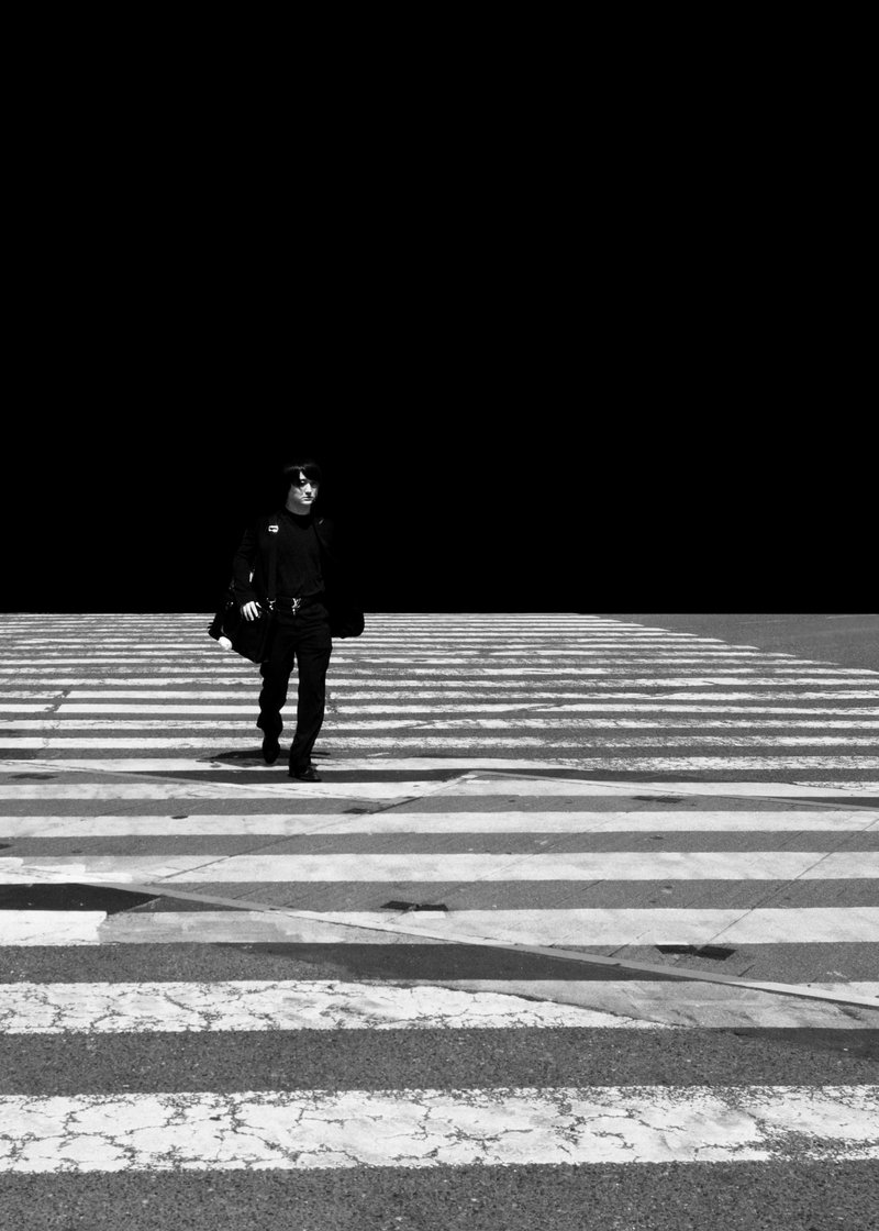 A person walking across a crosswalk in black and white, with a dark background and a few buildings faintly visible.
